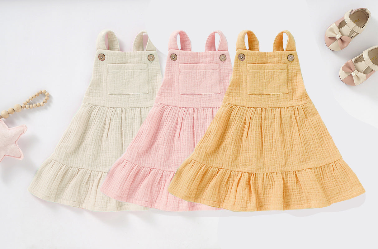 A soft and stylish Organic Cotton Muslin Baby Dress in three colors, featuring button straps and textured fabric, perfect for babies.