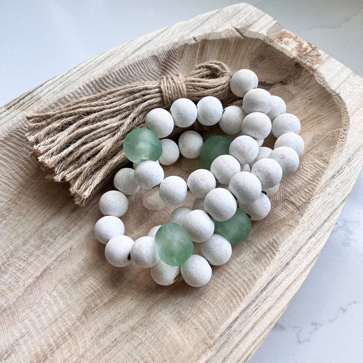 A beautifully handcrafted whitewashed wood bead garland featuring jumbo green and aqua recycled glass beads, adorned with jute tassels.