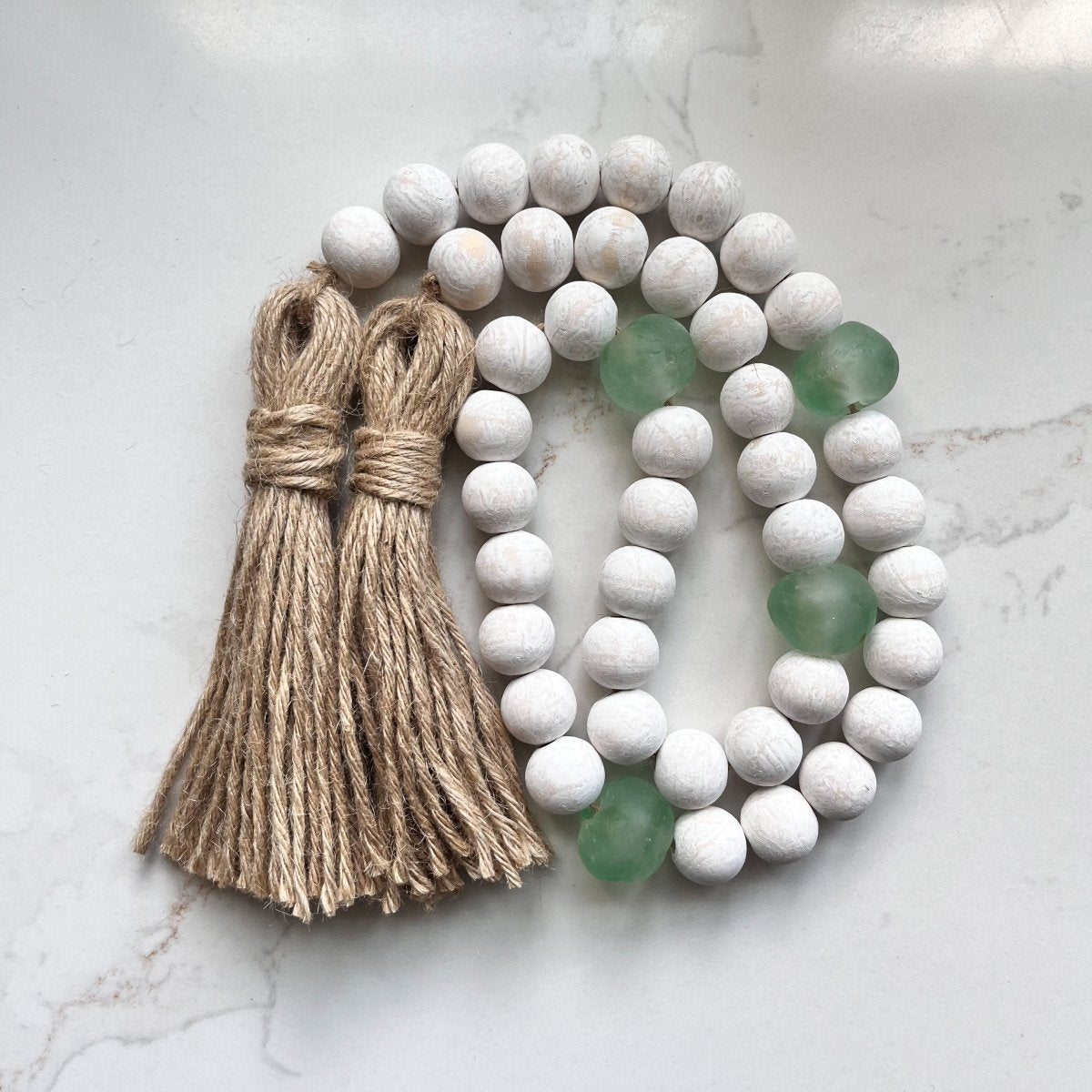 A beautifully handcrafted whitewashed wood bead garland featuring jumbo green and aqua recycled glass beads, adorned with jute tassels.