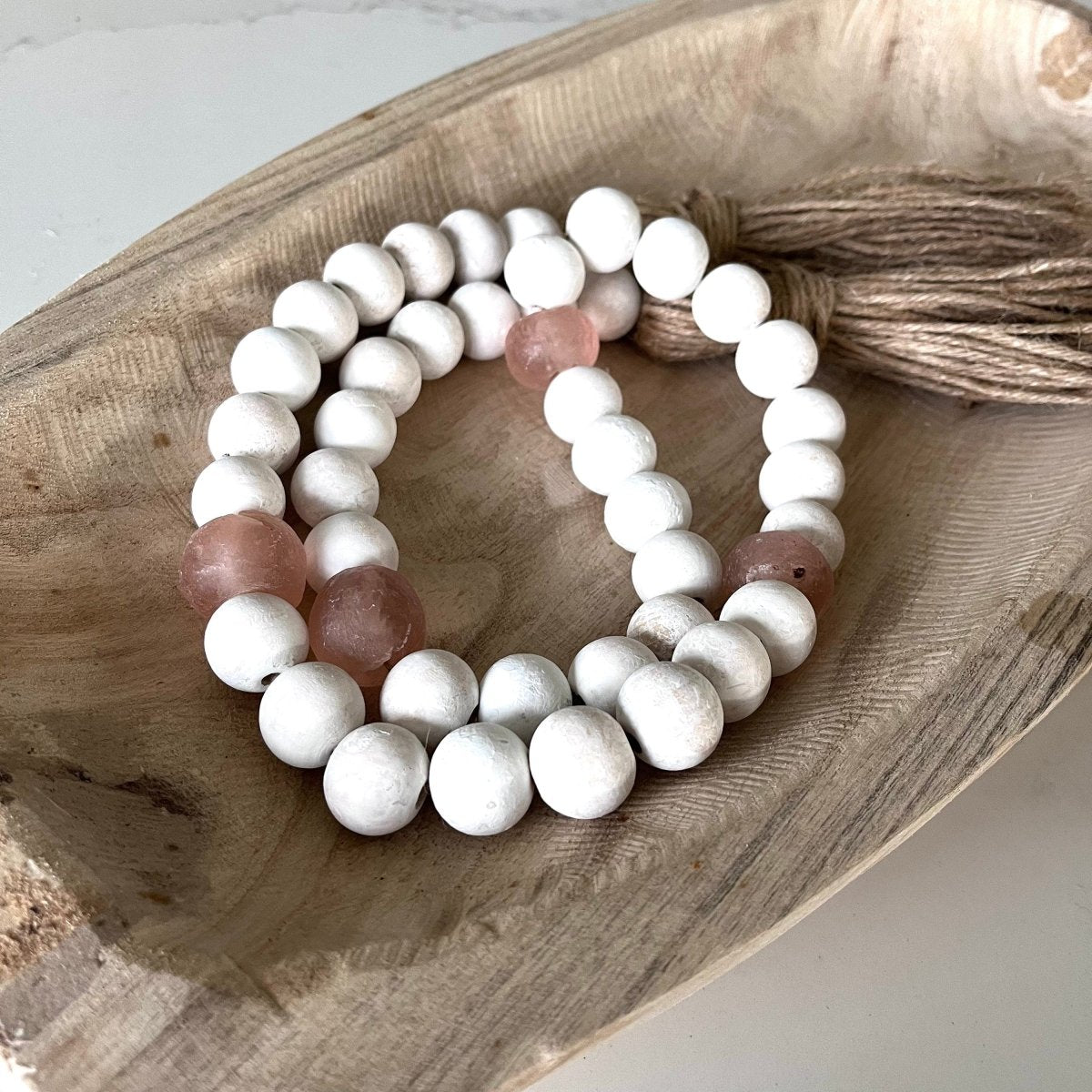 A beautiful whitewashed wood bead garland featuring jumbo rose recycled glass beads and jute tassels, perfect for coastal decor.