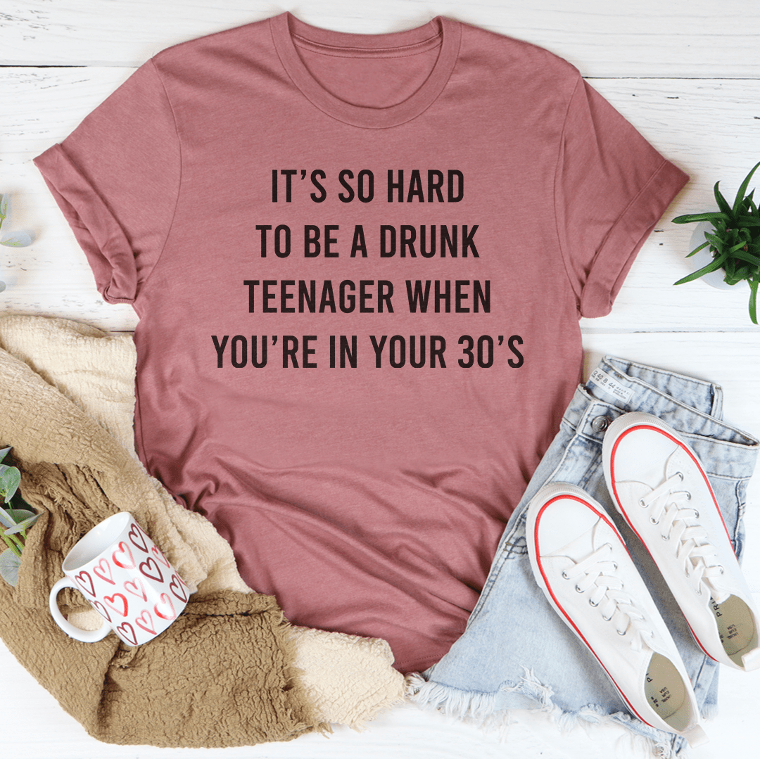A humorous T-shirt reading 'It's So Hard To Be A Drunk T-Shirtnager When You're In Yours 30's', made from soft cotton with double stitching.