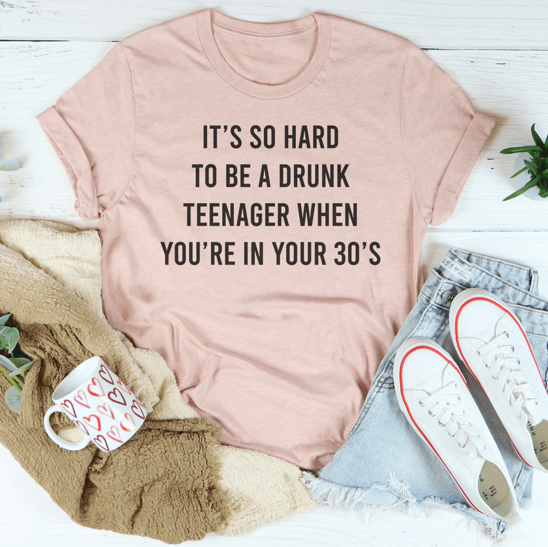 A humorous T-shirt reading 'It's So Hard To Be A Drunk T-Shirtnager When You're In Yours 30's', made from soft cotton with double stitching.