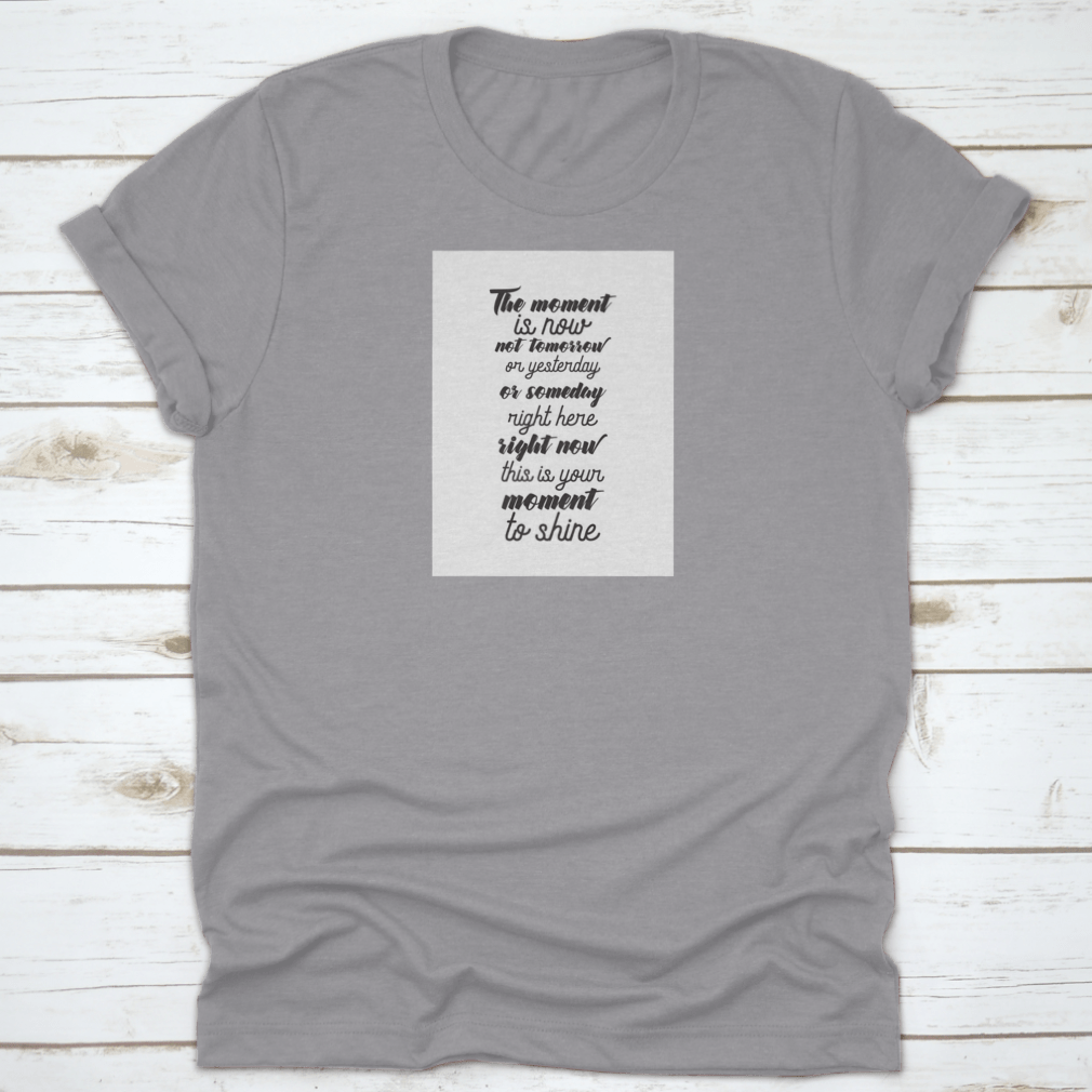 A motivational T-shirt featuring the quote 'The Moment Is Now, Not Tomorrow', made from soft cotton fabric, displayed on a neutral background.