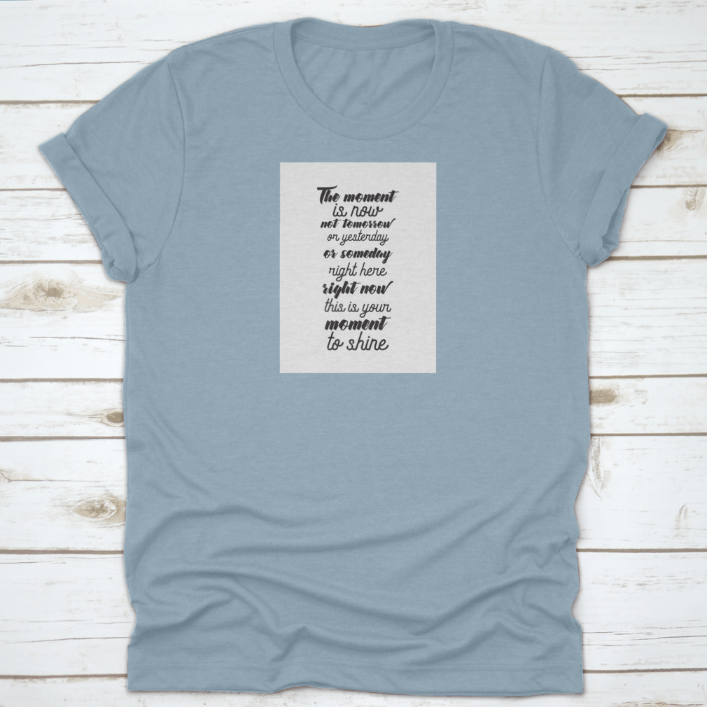 A motivational T-shirt featuring the quote 'The Moment Is Now, Not Tomorrow', made from soft cotton fabric, displayed on a neutral background.