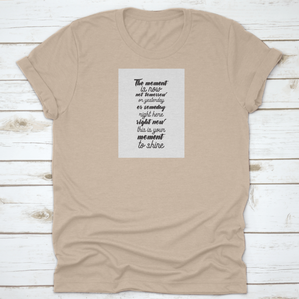 A motivational T-shirt featuring the quote 'The Moment Is Now, Not Tomorrow', made from soft cotton fabric, displayed on a neutral background.