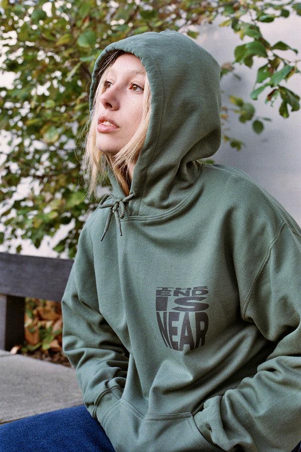 Military green hoodie featuring 'The End Is Near' printed design on the front, showcasing a relaxed fit and woven label details.