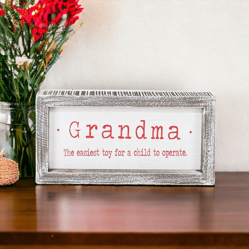 A humorous wood-framed sign that reads 'Grandma, The Easiest Toy For A Child To Operate' in white and red colors.