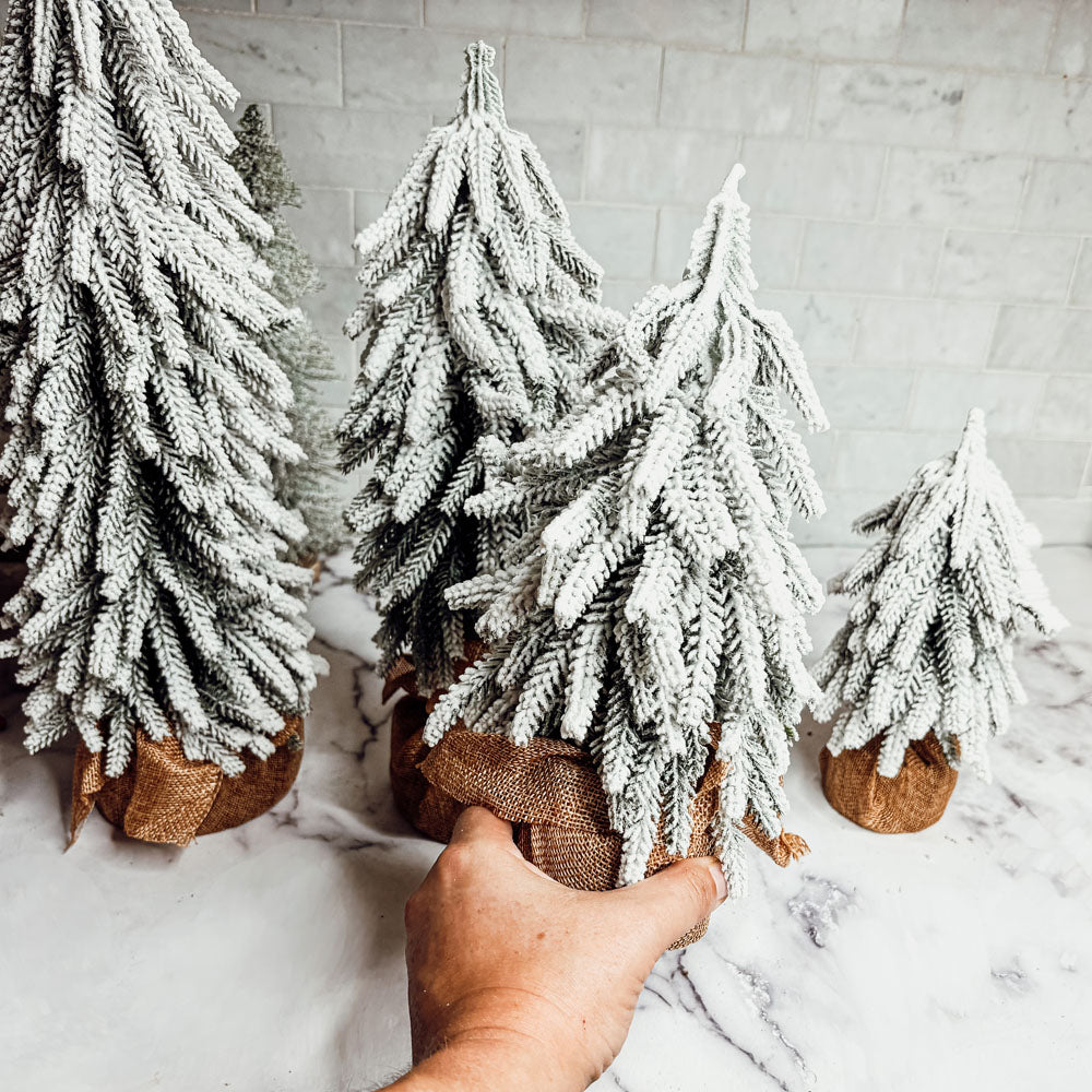 A set of four snow-covered Christmas trees in varying sizes, featuring a jute-covered wooden base, perfect for holiday decor.