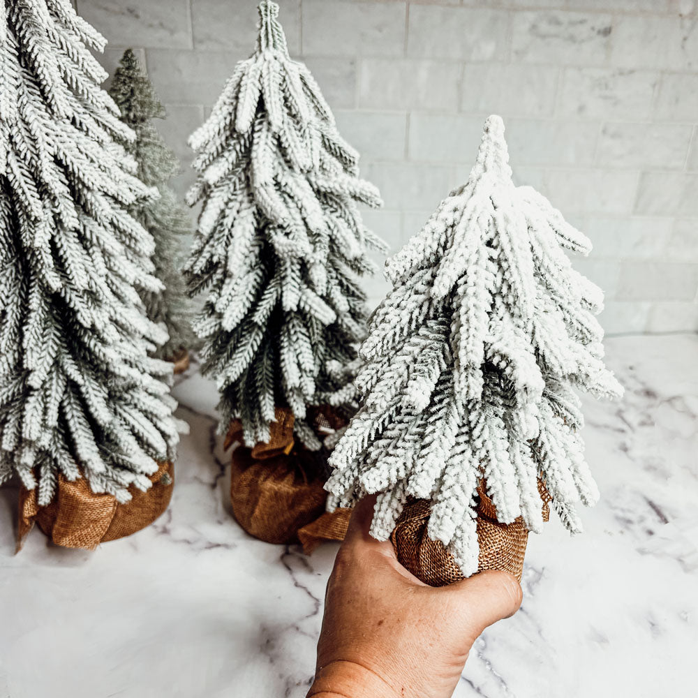 A set of four snow-covered Christmas trees in varying sizes, featuring a jute-covered wooden base, perfect for holiday decor.