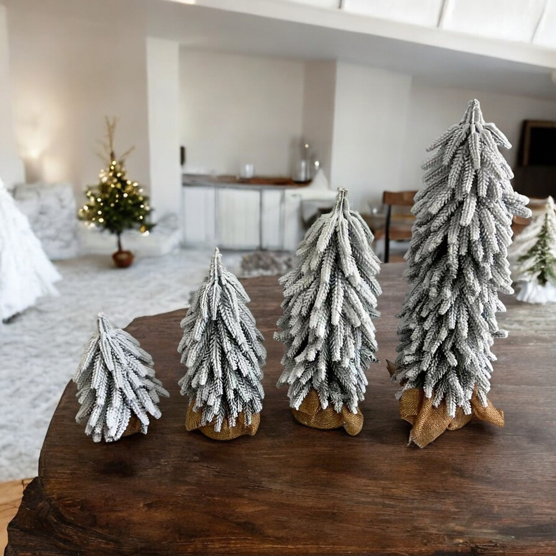 A set of four snow-covered Christmas trees in varying sizes, featuring a jute-covered wooden base, perfect for holiday decor.