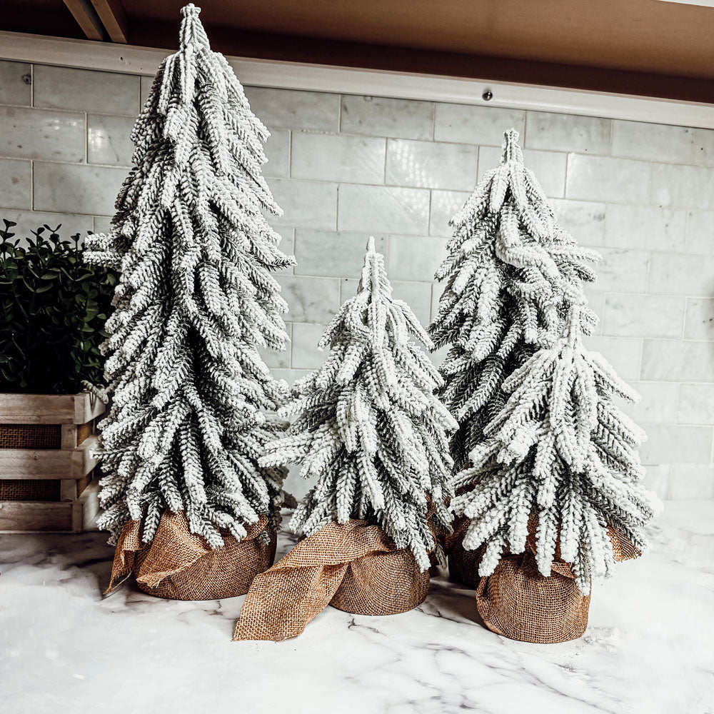 A set of four snow-covered Christmas trees in varying sizes, featuring a jute-covered wooden base, perfect for holiday decor.