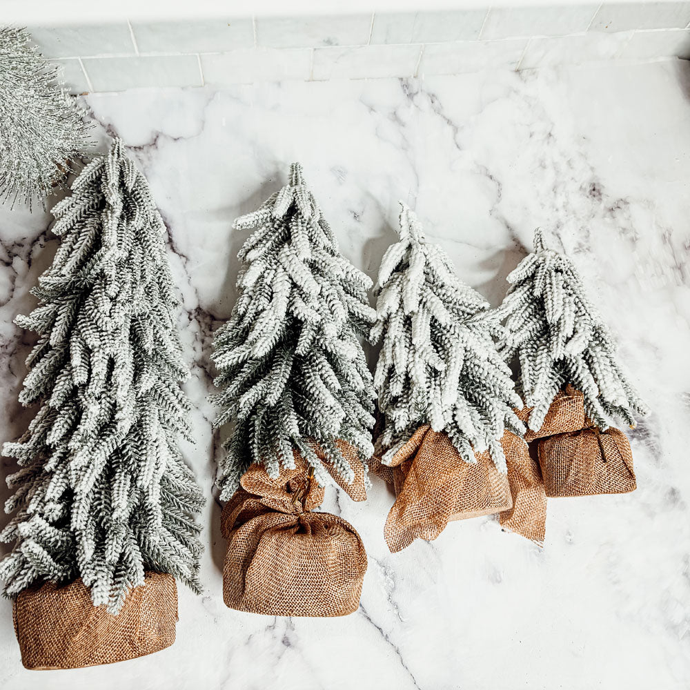 A set of four snow-covered Christmas trees in varying sizes, featuring a jute-covered wooden base, perfect for holiday decor.
