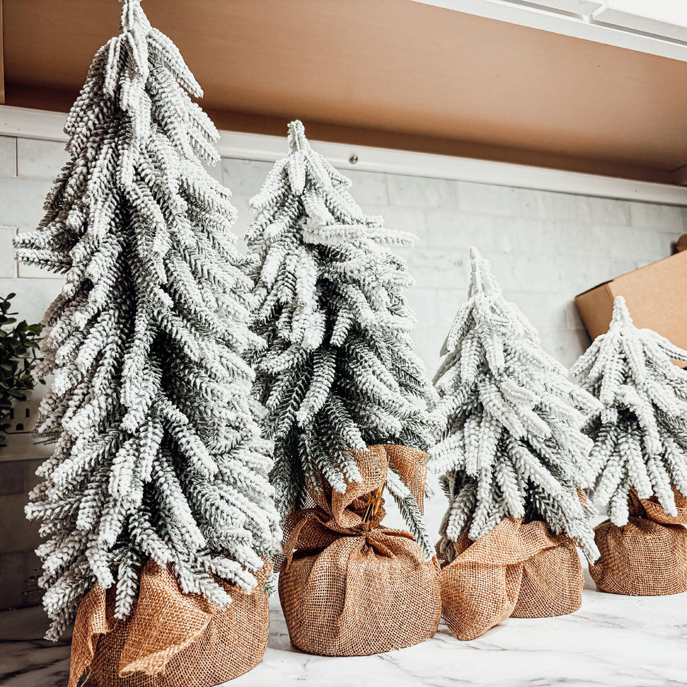 A set of four snow-covered Christmas trees in varying sizes, featuring a jute-covered wooden base, perfect for holiday decor.
