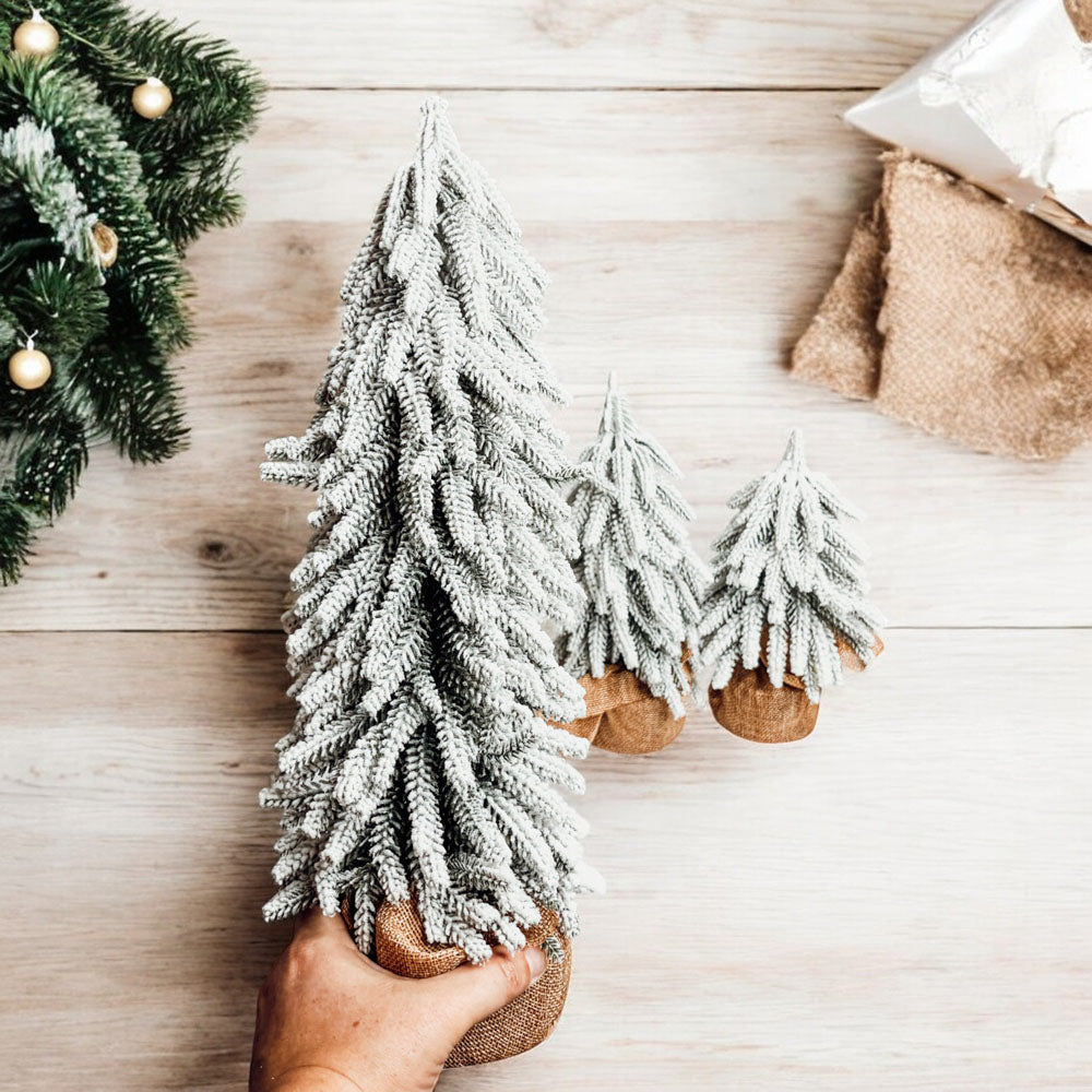 A set of four snow-covered Christmas trees in varying sizes, featuring a jute-covered wooden base, perfect for holiday decor.