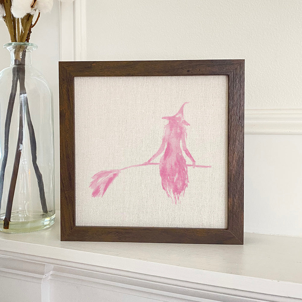 Framed sign featuring a pink witch silhouette on a linen-look background with a stylized wood frame.