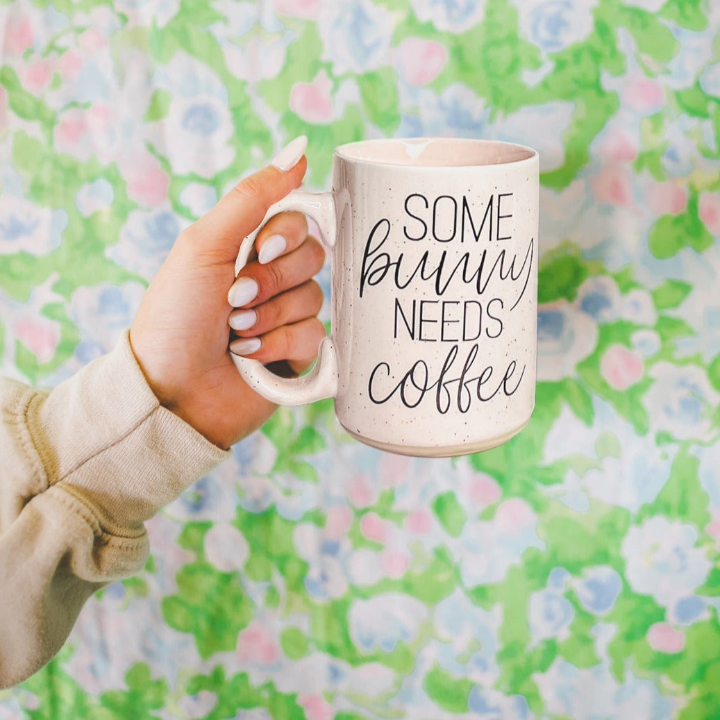 Ceramic coffee mug with pastel blue and pink colors, featuring the phrase 'Some Bunny Needs Coffee' on both sides, perfect for Easter and spring.
