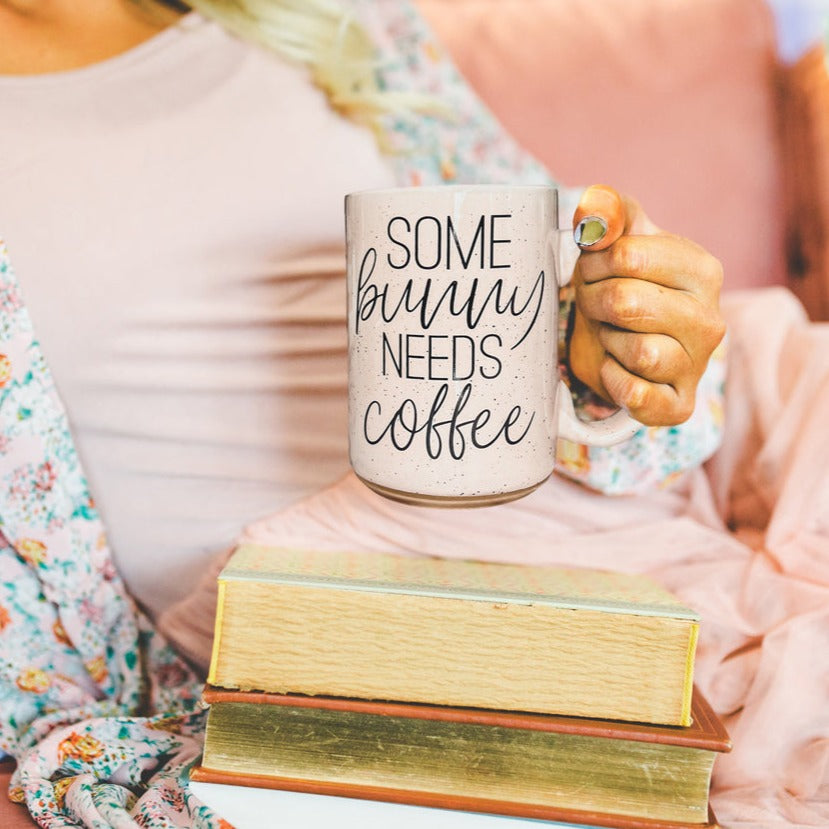 Ceramic coffee mug with pastel blue and pink colors, featuring the phrase 'Some Bunny Needs Coffee' on both sides, perfect for Easter and spring.