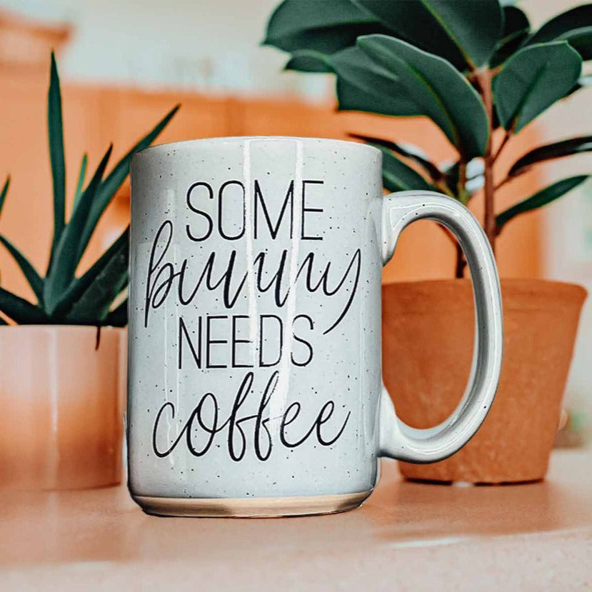 Ceramic coffee mug with pastel blue and pink colors, featuring the phrase 'Some Bunny Needs Coffee' on both sides, perfect for Easter and spring.