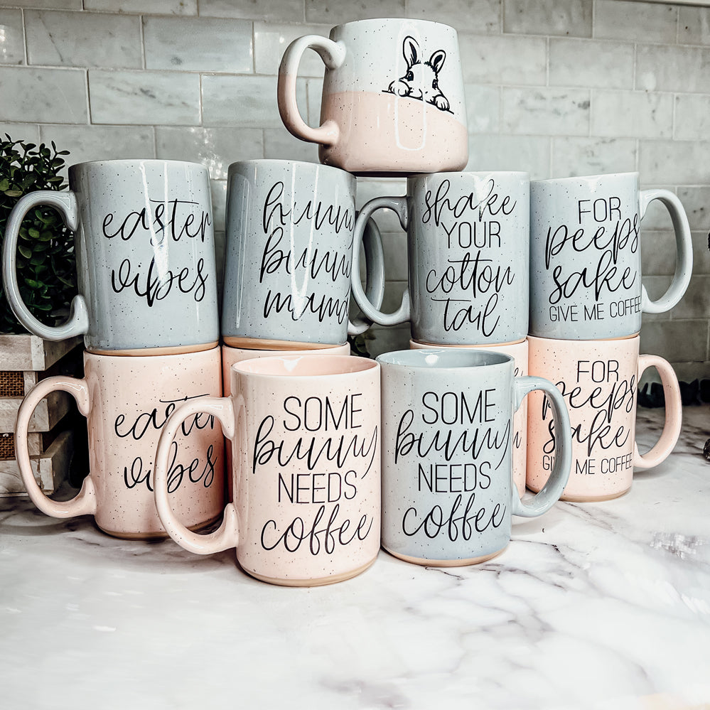 Ceramic coffee mug with pastel blue and pink colors, featuring the phrase 'Some Bunny Needs Coffee' on both sides, perfect for Easter and spring.