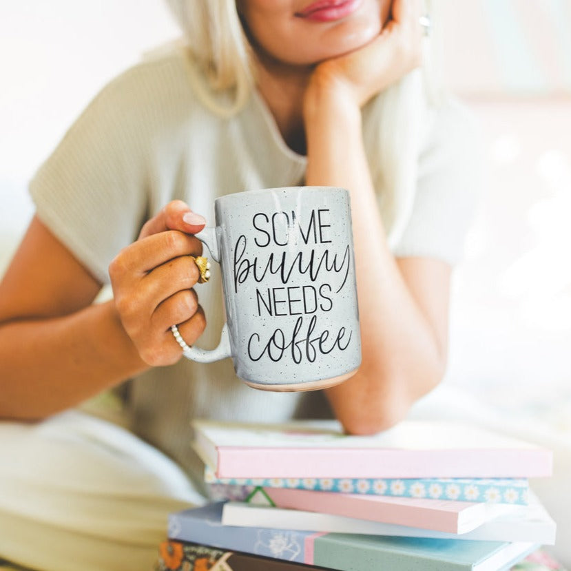 Ceramic coffee mug with pastel blue and pink colors, featuring the phrase 'Some Bunny Needs Coffee' on both sides, perfect for Easter and spring.