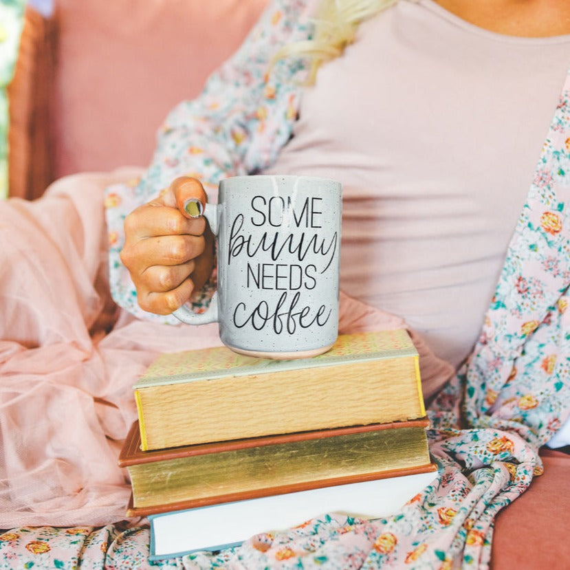 Ceramic coffee mug with pastel blue and pink colors, featuring the phrase 'Some Bunny Needs Coffee' on both sides, perfect for Easter and spring.