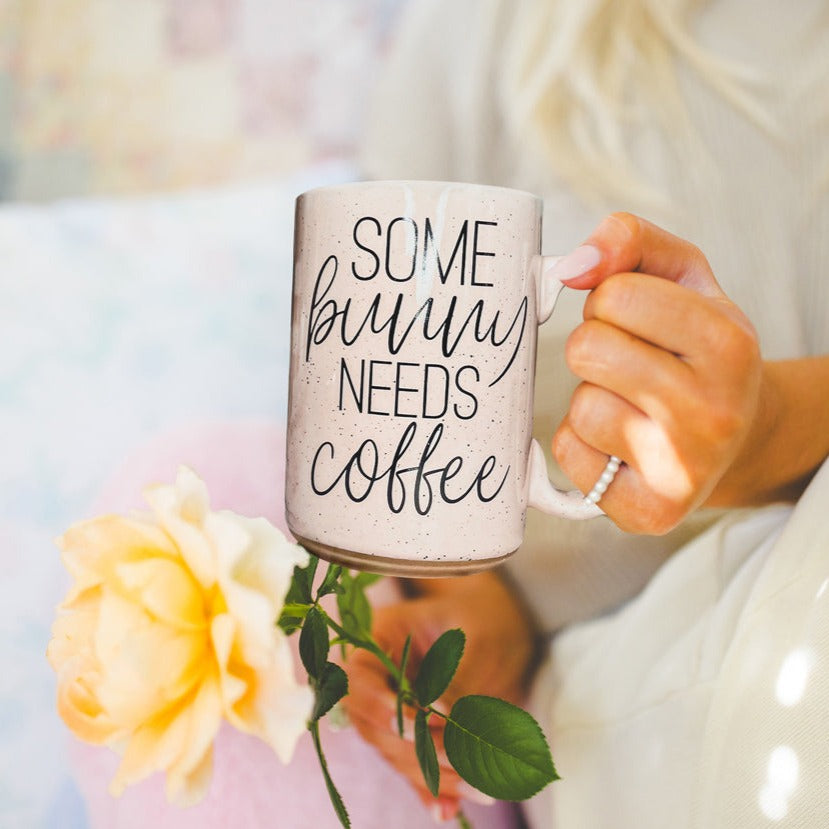 Ceramic coffee mug with pastel blue and pink colors, featuring the phrase 'Some Bunny Needs Coffee' on both sides, perfect for Easter and spring.