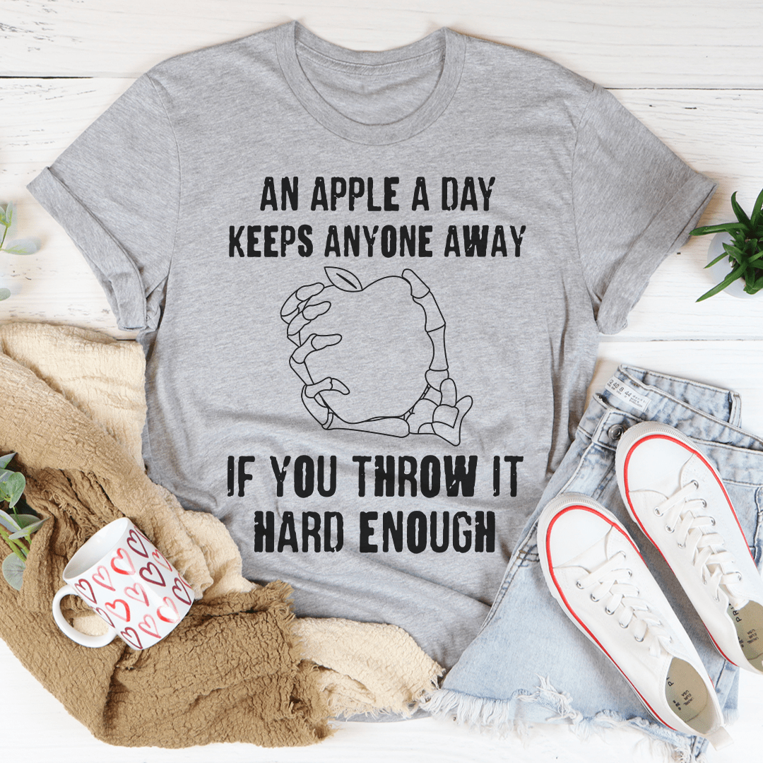 A humorous t-shirt featuring the phrase 'An Apple A Day Keeps Anyone Away If You Throw It Hard Enough' printed on soft cotton fabric.