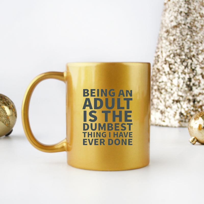 Ceramic mug with gold and silver design featuring the phrase 'Being An Adult Is The Dumbest Thing I Have Ever Done'.