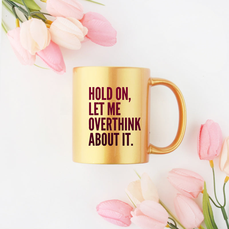 Elegant gold and silver ceramic mug with the phrase 'Hold On Let Me Overthink About It' printed on it, showcasing a stylish design.