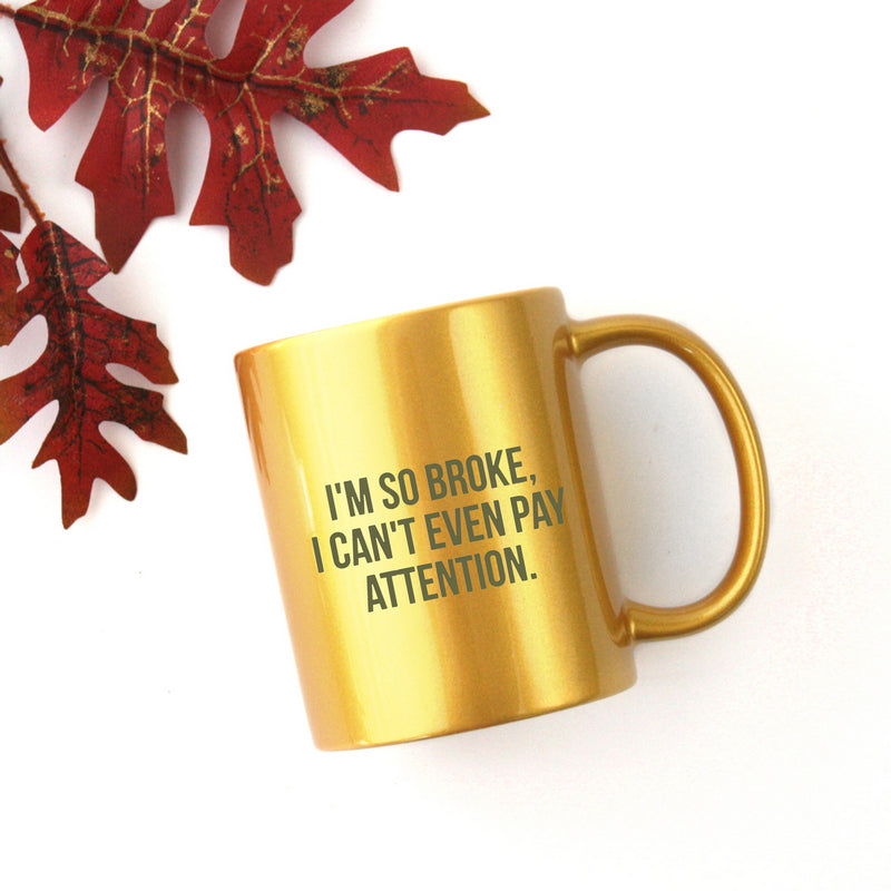 Ceramic mug with gold metallic coating featuring humorous text 'I'm So Broke I Can't Even Pay Attention' on a white background.