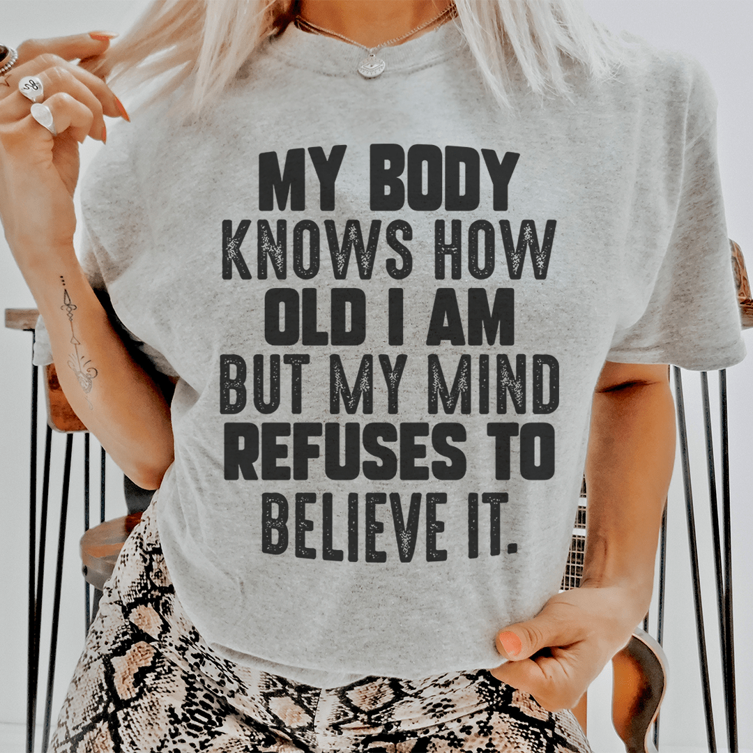 A stylish t-shirt featuring the phrase 'My Body Knows How Old I Am But My Mind Refuses To Believe It', made from soft cotton with double stitching.