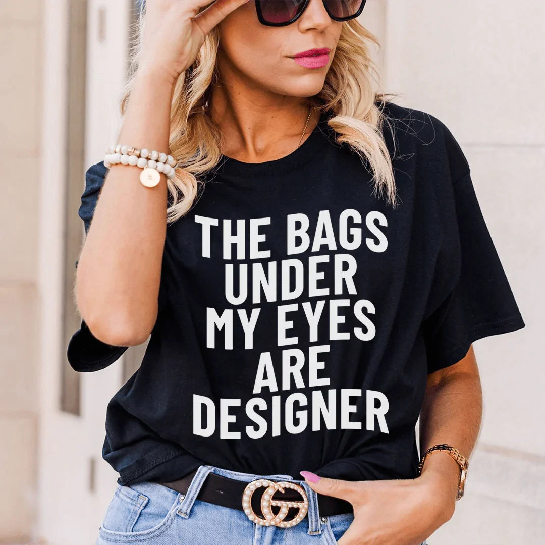 A stylish t-shirt featuring the phrase 'The Bags Under My Eyes Are Designers' printed on it, made from soft ring-spun cotton.