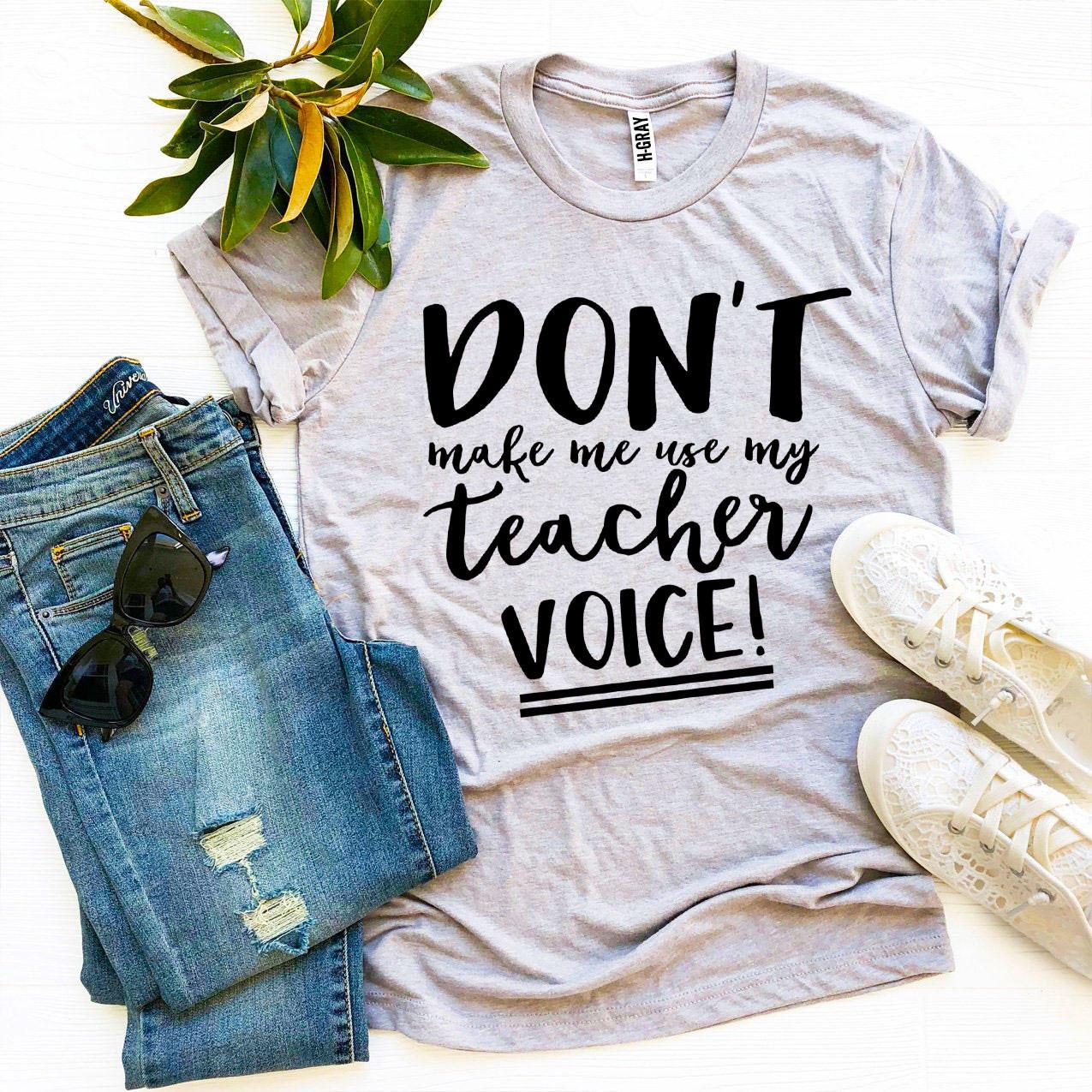 A black t-shirt featuring the phrase 'Don’t Make Me Use My Teacher Voice!' in bold white letters, made from soft ring spun cotton.