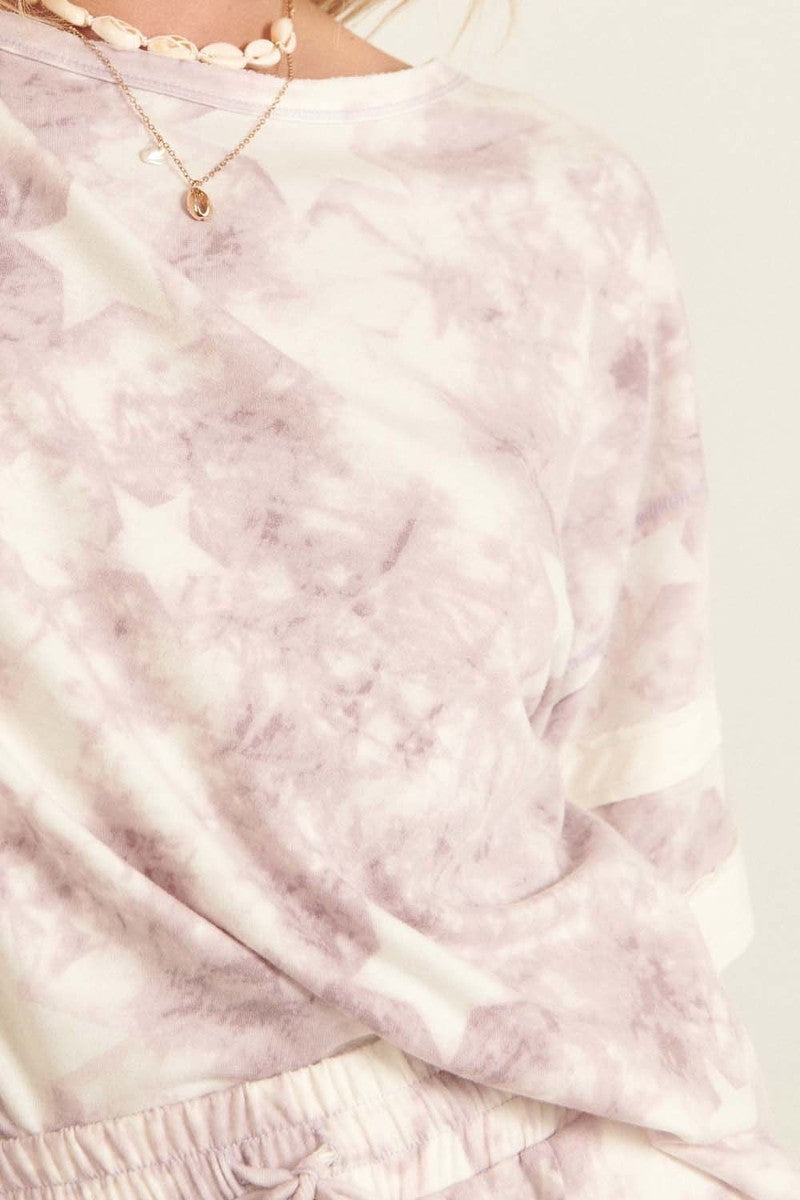 A lavender star-pattern tie-dye knit top with short sleeves and varsity stripe trim, showcasing a relaxed oversized fit.