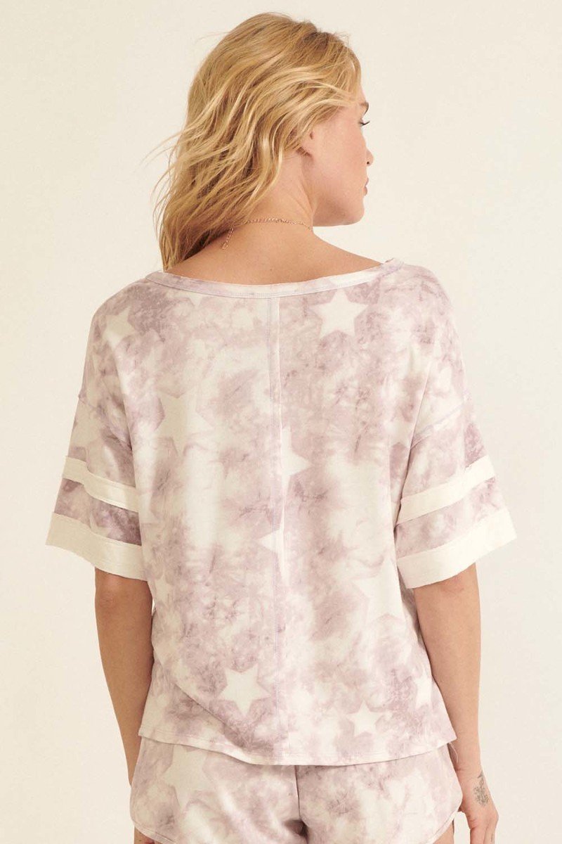 A lavender star-pattern tie-dye knit top with short sleeves and varsity stripe trim, showcasing a relaxed oversized fit.