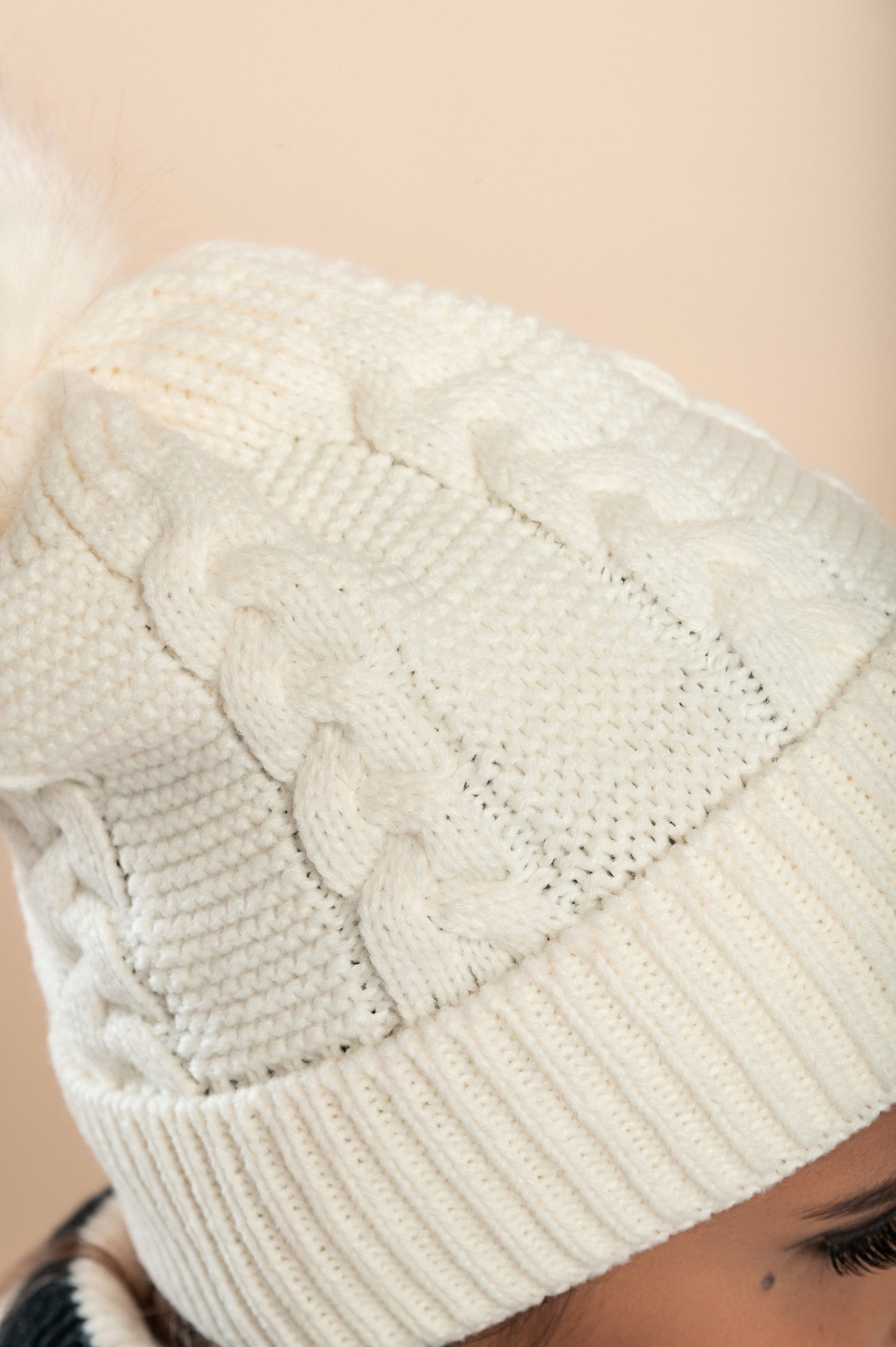 A stylish white knitted hat with a faux fur pompom on top, featuring a rolled edge and ribbed knit design.
