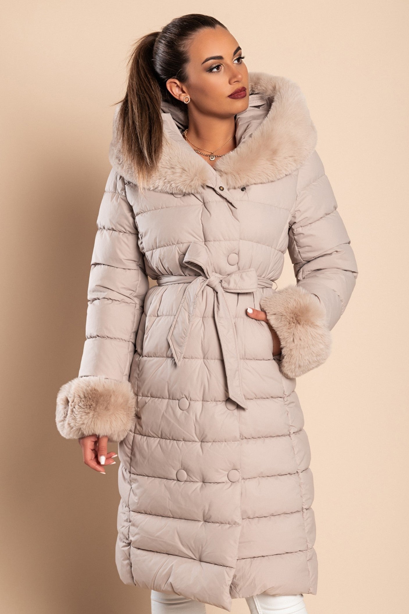 Long padded winter jacket in beige with removable faux fur collar, side pockets, and decorative belt, perfect for winter wear.