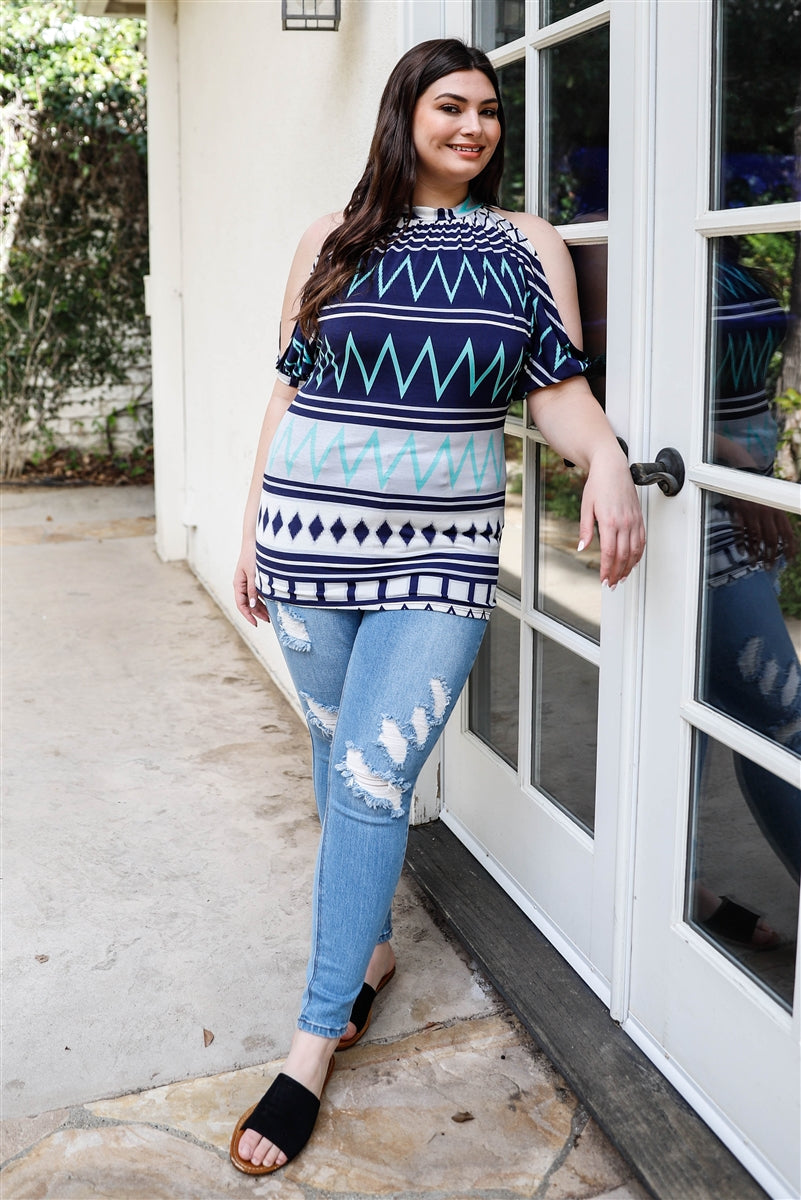 A stylish plus-size woman wearing a navy and mint polka dot cold shoulder top with short sleeves and a back self-tie detail.