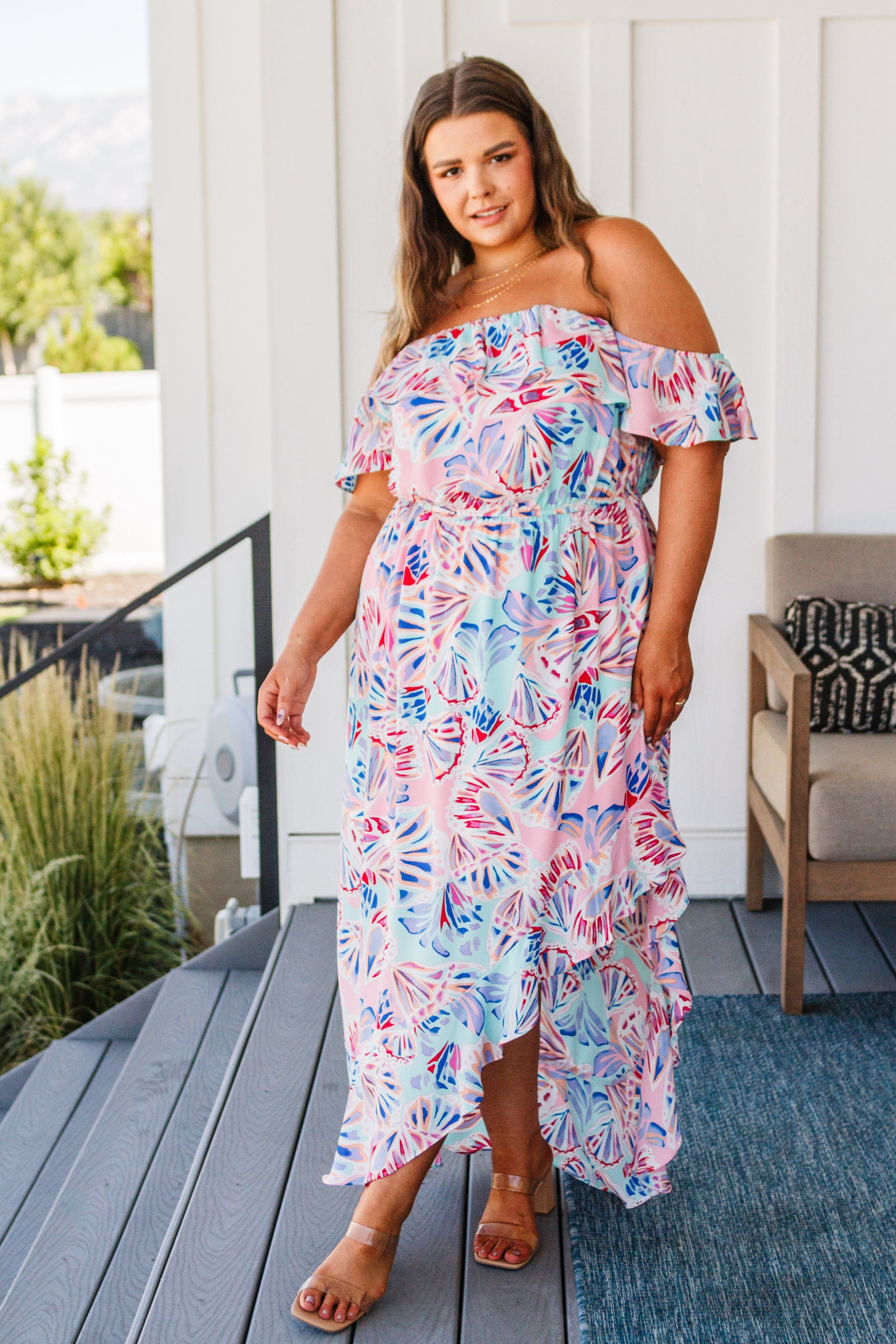 A beautiful model wearing the She Sells Sea Shells Maxi Dress featuring a ruffled neckline and asymmetrical hem in a breezy shell print.