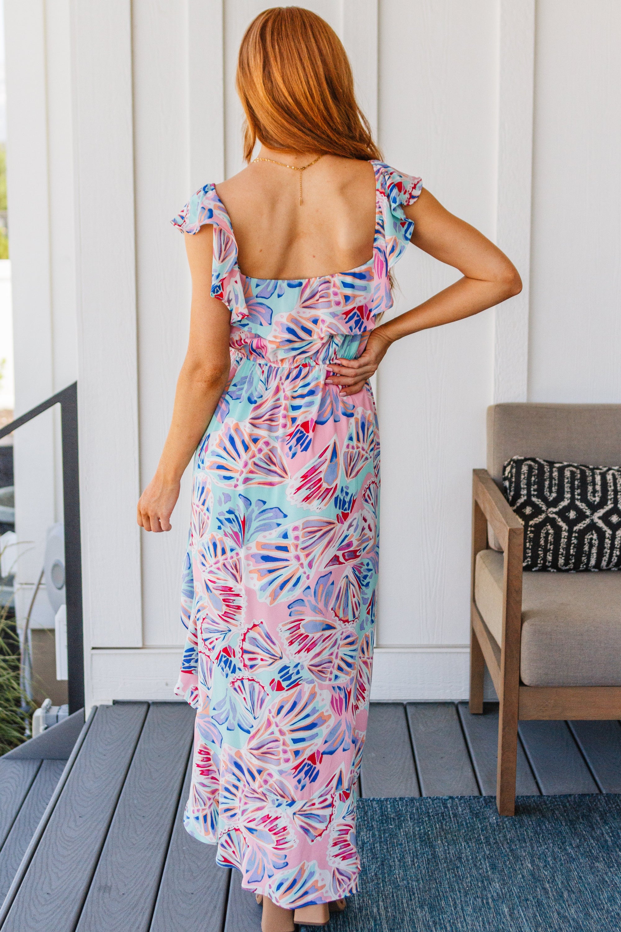 A beautiful model wearing the She Sells Sea Shells Maxi Dress featuring a ruffled neckline and asymmetrical hem in a breezy shell print.
