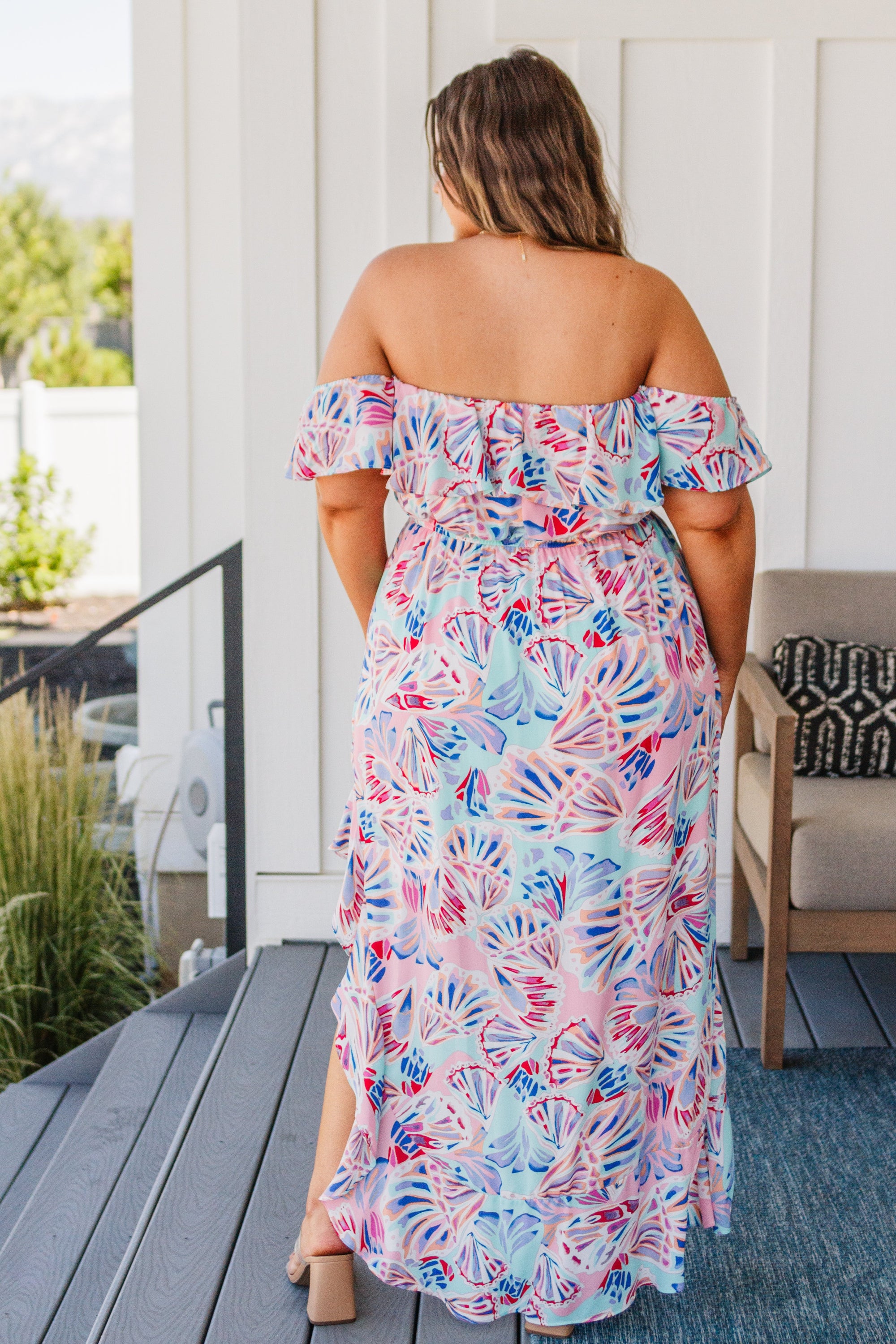 A beautiful model wearing the She Sells Sea Shells Maxi Dress featuring a ruffled neckline and asymmetrical hem in a breezy shell print.